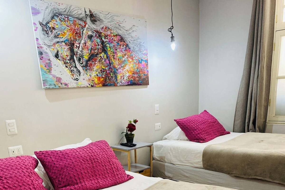 Hotelkamer met twee bedden, elk met een beige dekbedovertrek en roze kussens. Er hangt een schilderij aan de muur, een klein tafeltje met een bloempot en een lamp.