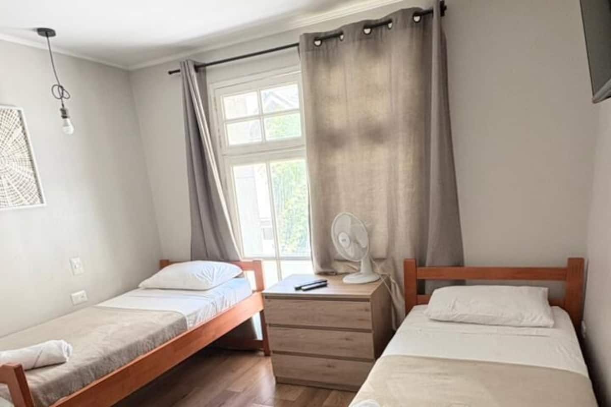 Een kamer met twee houten bedden, een tafeltje, een ventilator en een raam met gordijnen.