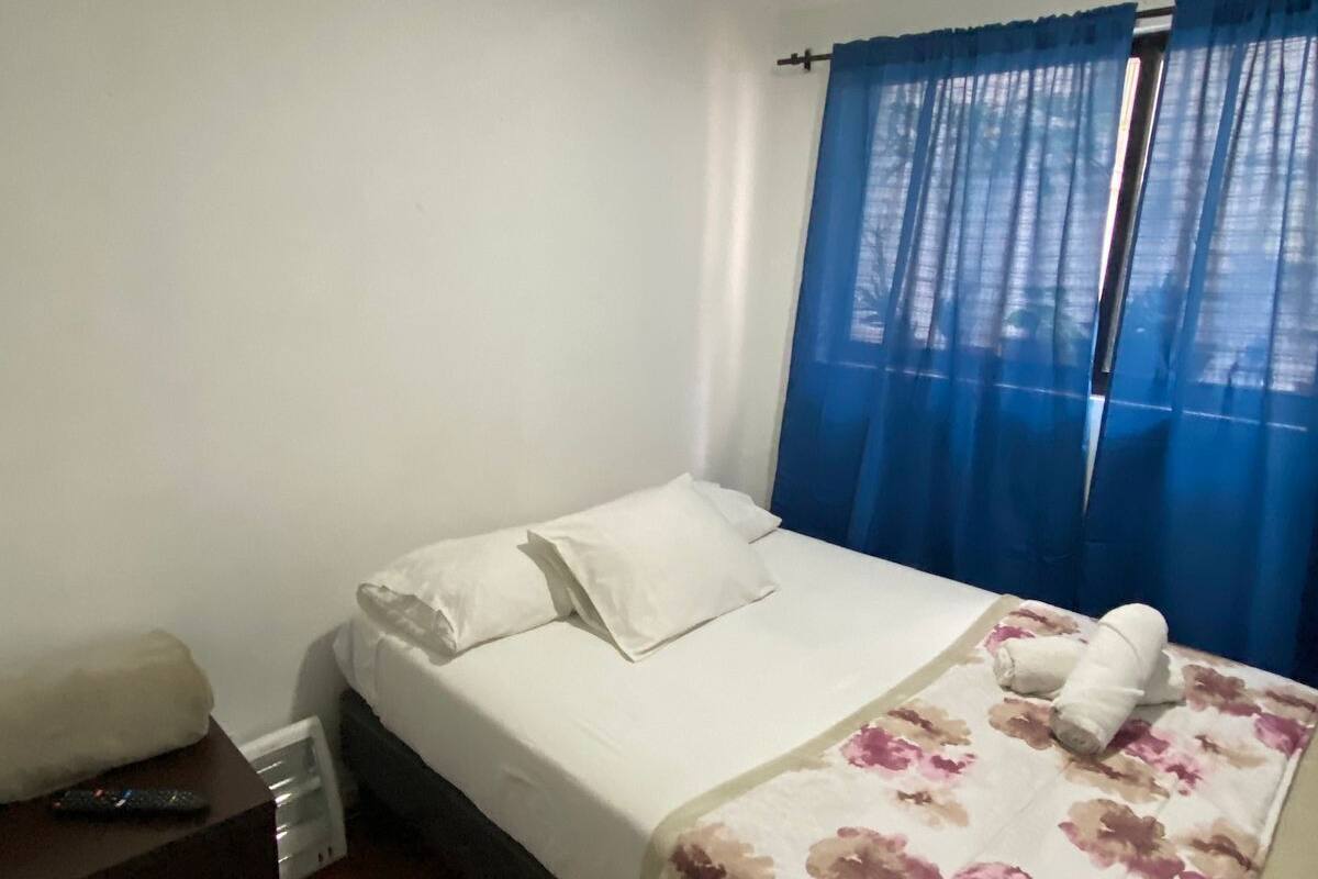 Een eenpersoonsbed met een bloemenprint sprei, witte kussens en een afstandsbediening op een klein tafeltje.