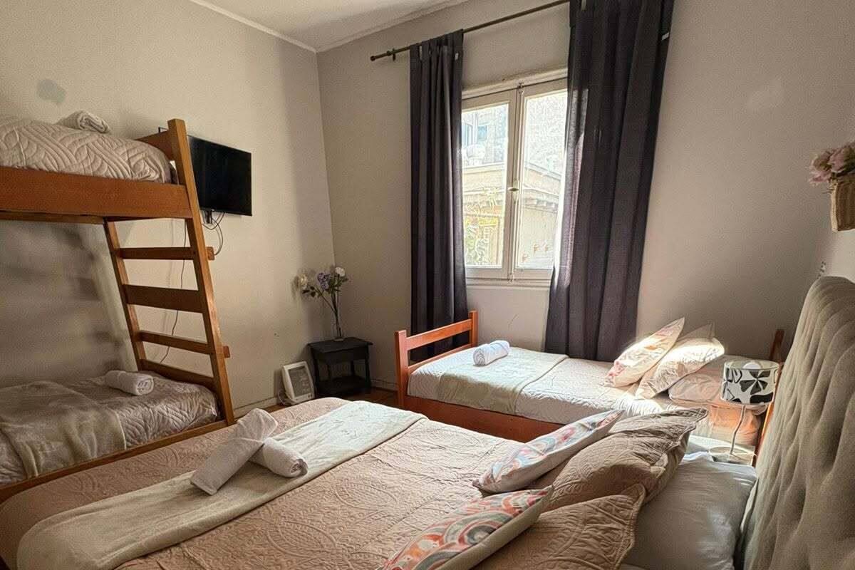 Een kamer met twee bedden, een stapelbed, een bureau met lamp, een raam met gordijnen, en een kleine tafel met een vaas.