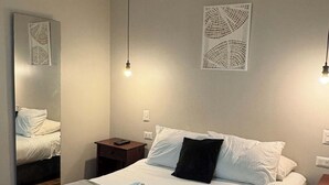 Un lit bien fait, avec du linge de lit blanc et un oreiller noir, une table de chevet avec une télécommande et un luminaire fixé au mur.