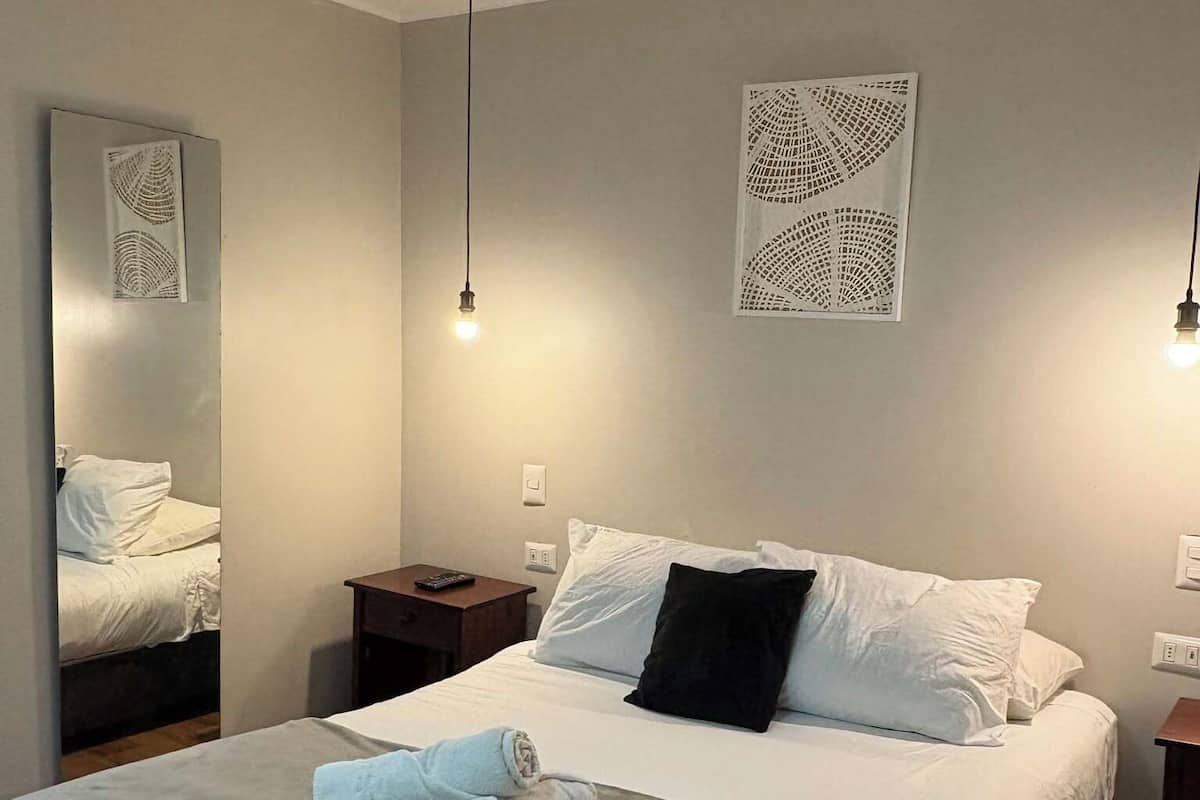 Een keurig opgemaakt bed met witte lakens en een zwart kussen, een nachtkastje met een afstandsbediening, en een wandlamp.