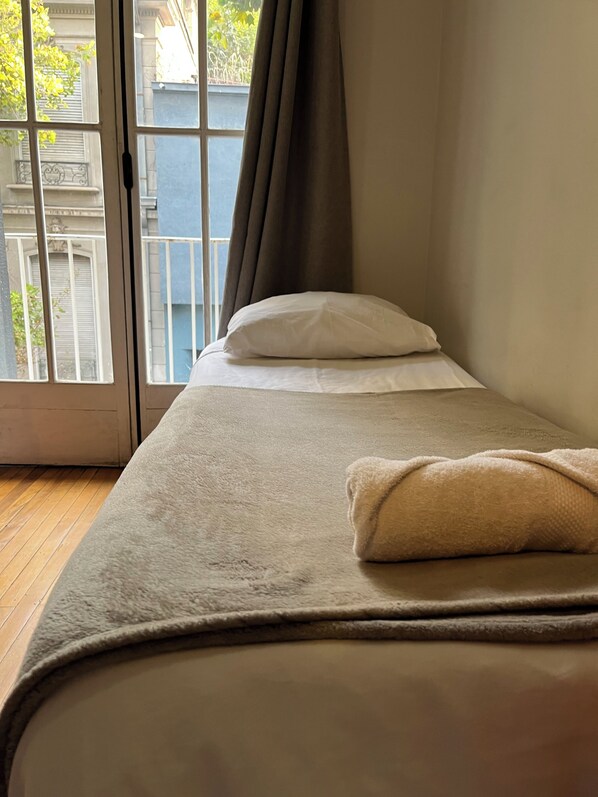 Un lit simple avec un oreiller blanc et une couverture beige, placé à côté d’une porte-fenêtre coulissante donnant sur un espace extérieur avec un mur bleu et de la verdure.