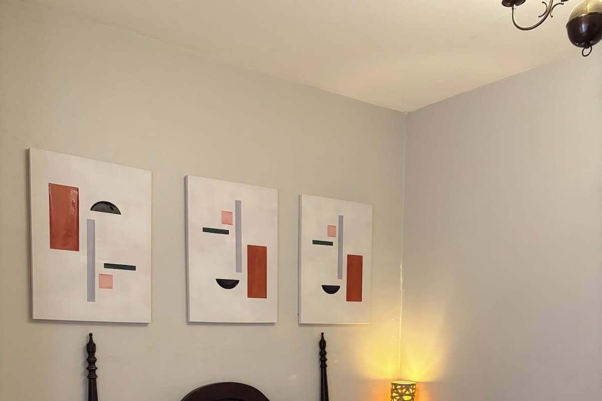 Een net opgemaakt bed met een grijze plaid, twee kussens en een opgerolde handdoek ernaast. Boven het bed hangen drie abstracte schilderijen aan de muur.