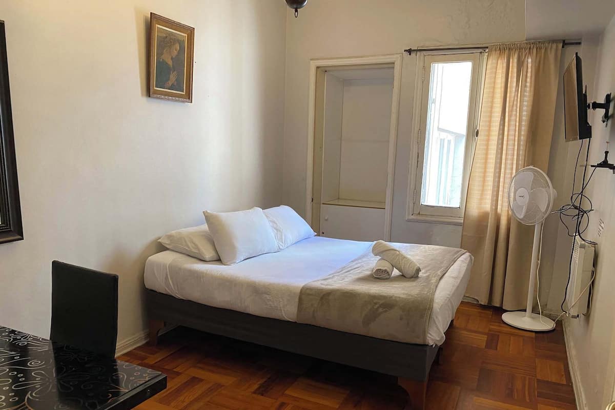 Een slaapkamer met een bed, een bureau, een stoel, een foto aan de muur, een raam met gordijnen en een staande ventilator.