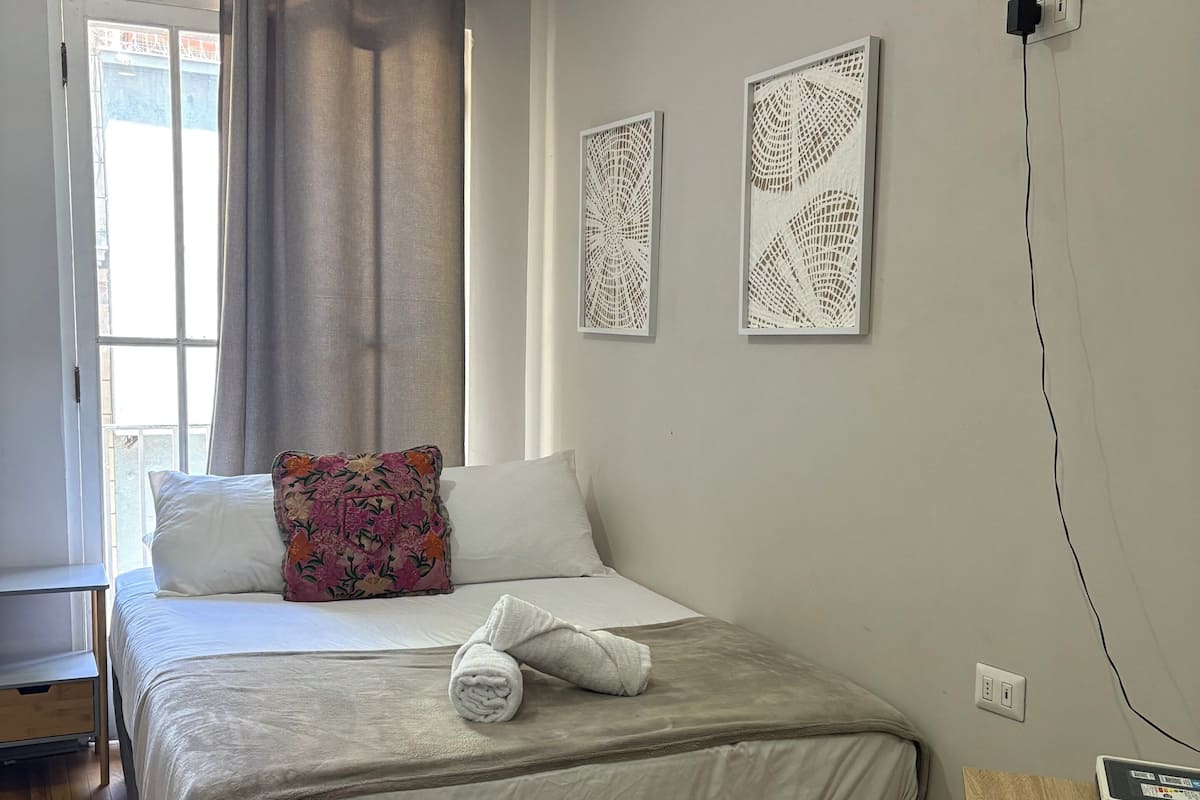 Een slaapkamer met een bed, een raam met gordijnen, een nachtkastje en twee decoratieve items aan de muur.