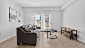 A spacious living room with a grey sofa, a wooden coffee table, and a bench. There is a large window with blinds, a door leading outside, and a wall-mounted artwork. - Lux Studio ,Pool, Near St. Davids Hospital (Jarrell)