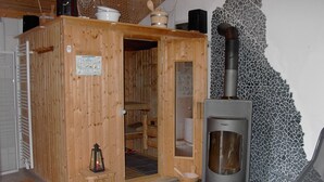 Uma sala de sauna com estrutura de madeira, porta de vidro, chão ladrilhado, um banquinho, um candeeiro e um forno para sauna.