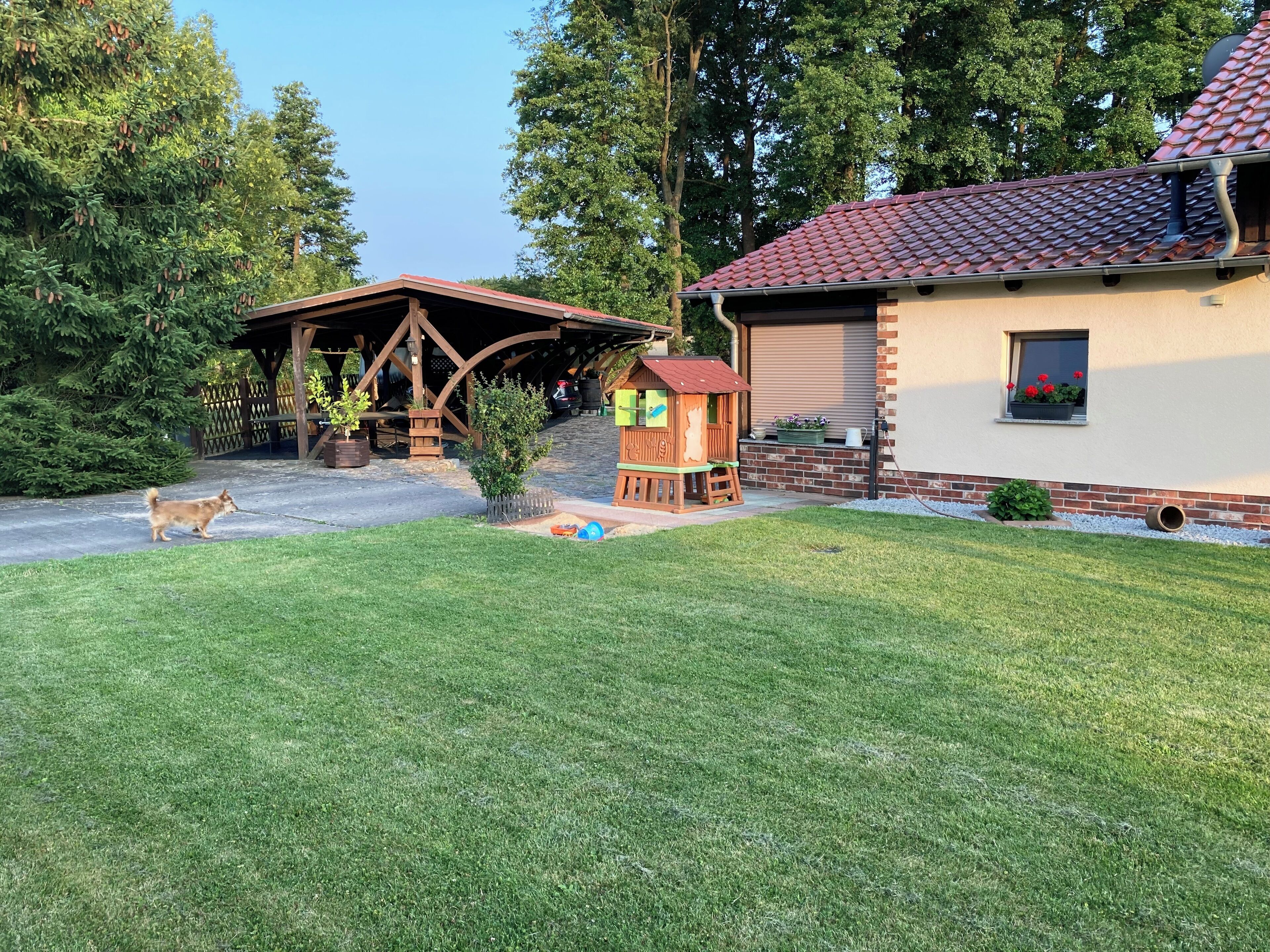Une maison avec un toit recouvert de tuiles rouges, une cabane en bois pour jouer et un jardin clos.