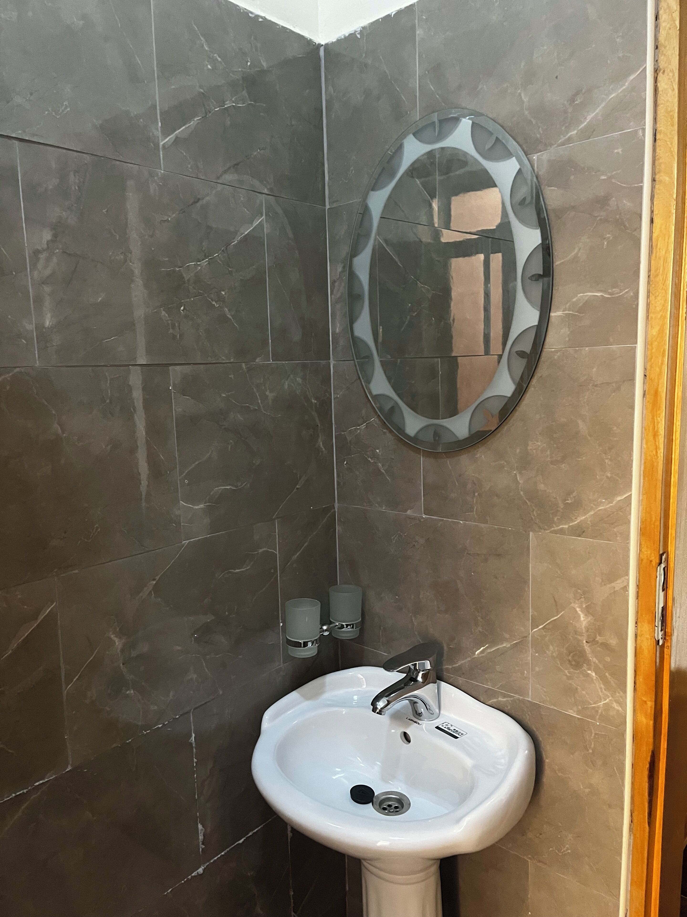 A bathroom with dark marble tiles, a white sink, a mirror with a circular frame, and two wall-mounted soap dispensers.