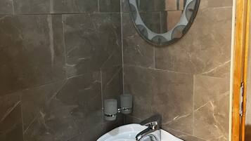 A bathroom with dark marble tiles, a white sink, a mirror with a circular frame, and two wall-mounted soap dispensers.