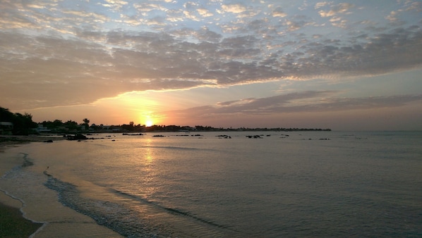 A beach with the sun setting over the horizon, calm water, and a distant shoreline with buildings.