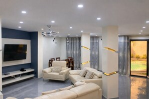 A modern living room with a flat-screen TV, leather sofas, a dining table, and a ceiling light fixture.