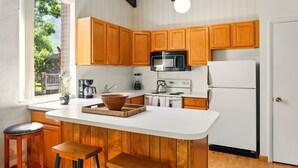 A kitchen with wooden cabinets, a white countertop, a sink, a stove, a microwave, and a refrigerator. There are two stools and a window with a view of greenery outside. - Cozy 2BR Ogden Condo - Pool, Trails, Snowbasin (Ogden)