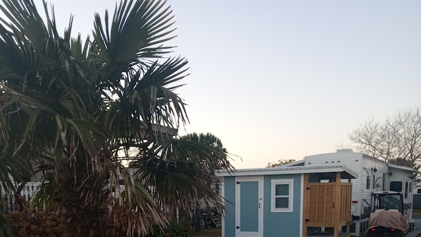 A small blue mobile home with a wooden deck and a fire pit in the front yard.