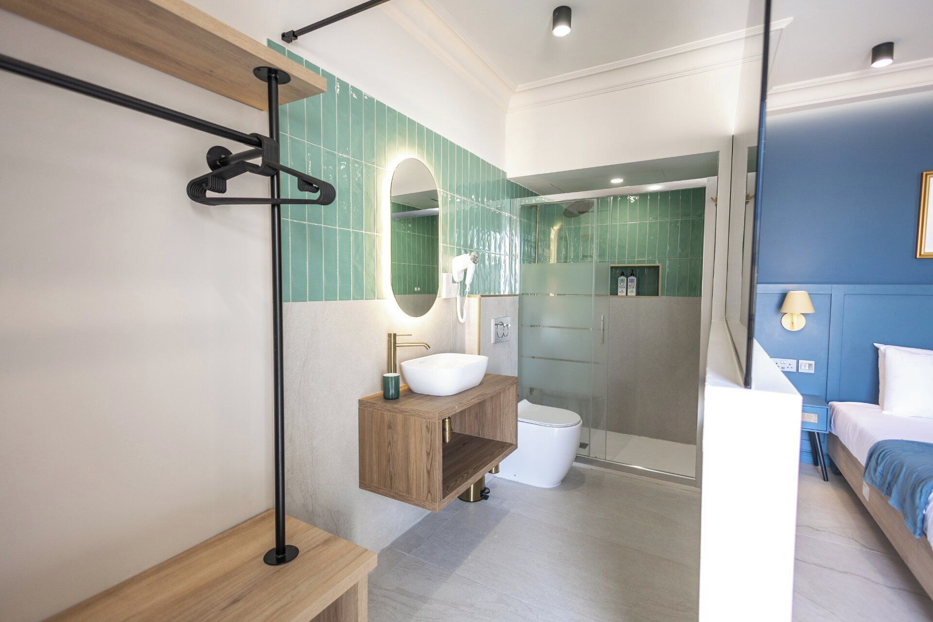 A modern bathroom with a wooden vanity, a freestanding mirror, a glass shower enclosure, and a bed in the adjacent room.