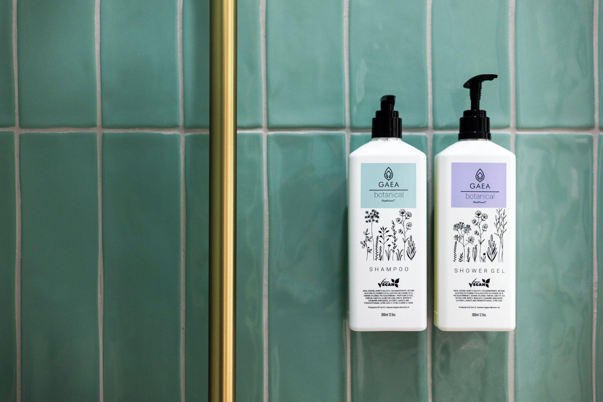 Two bottles of Gaea botanical shampoo and shower gel against a green tiled wall.