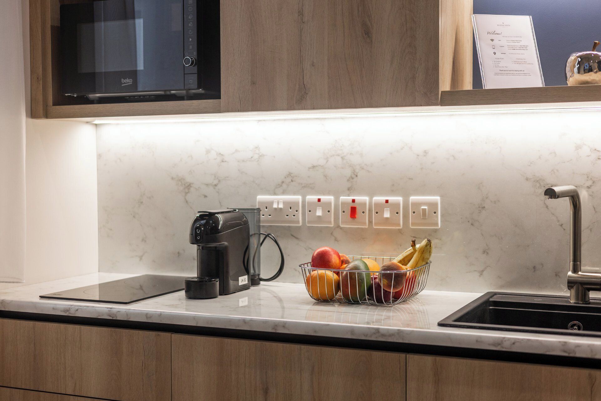 A kitchen with a built-in microwave, a coffee maker, a fruit basket, and a sink.