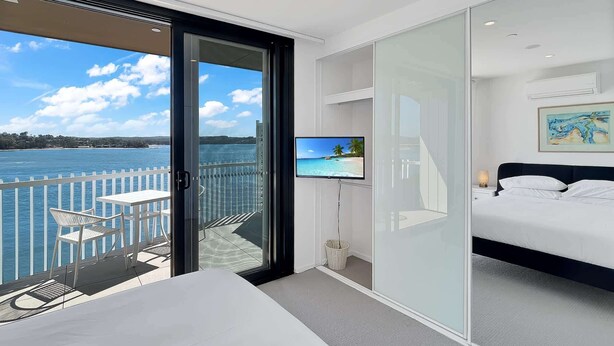 A modern hotel room with a balcony overlooking a body of water, a bed, a TV, and a painting on the wall.
