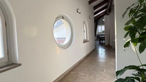 Un couloir doté d’un plancher poli, de murs blancs et de poutres en bois. On y trouve trois fenêtres en arc de cercle et, sur la droite, un pot de fleurs.