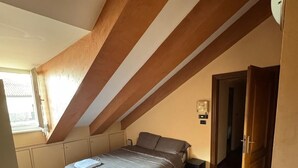 Une chambre avec un plafond en pente, un plancher en bois, un lit recouvert d’une housse de lit grise et une fenêtre laissant entrer la lumière naturelle.