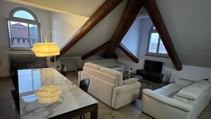 Un appartement-loft spacieux avec de hauts plafonds, des poutres apparentes, une table à manger avec des chaises, un canapé et un téléviseur.