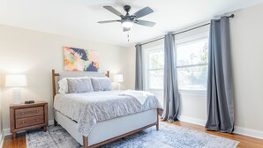 Une chambre à coucher comprenant un lit, des tables de chevet, un ventilateur de plafond, une fenêtre avec des rideaux et un tableau accroché au mur.