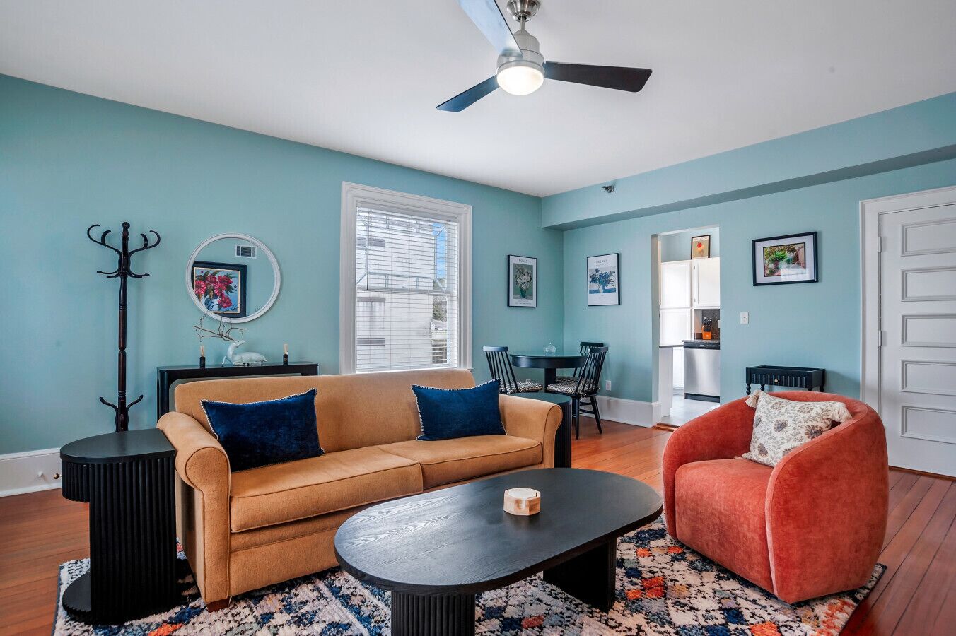 A living room with a sofa, a coffee table, a red armchair, a dining table with chairs, a kitchen area, and a coat rack.