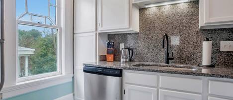 A kitchen with white cabinets, a granite countertop, a dishwasher, a sink with a black faucet, and a window with a view of trees.