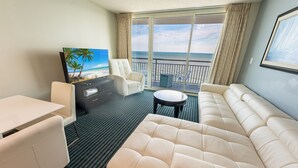 Habitación de hotel con una ventana grande que ofrece vistas a la playa, un sofá, una mesa de comedor y un televisor.