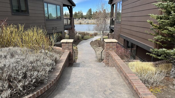 A pathway leading to a building with a view of a lake, surrounded by shrubs and a small tree. - Beautiful end unit home with big horizon views! (Bend)