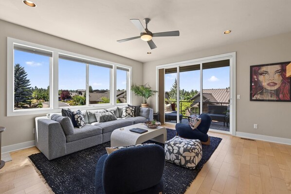 A spacious living room with a grey sectional sofa, a coffee table, and a blue armchair. There is a ceiling fan, large windows, and a view of the outdoors. - Beautiful end unit home with big horizon views! (Bend)