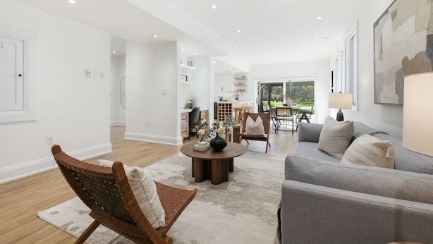 A spacious living room with a grey sofa, a wooden chair, and a coffee table. There is a dining area with a table and chairs, and a kitchen in the background. - Lake Simcoe Ice Fishing Basecamp • Sleeps 10–12 • Heated Shop (Beaverton)