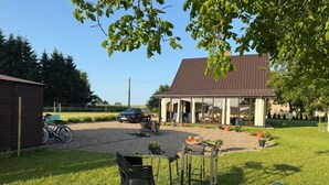 Ein überdachter Außenbereich mit Tisch und Stühlen, eine Schotterauffahrt, ein abgestelltes Fahrrad und ein Gebäude mit geneigtem Dach im Hintergrund.