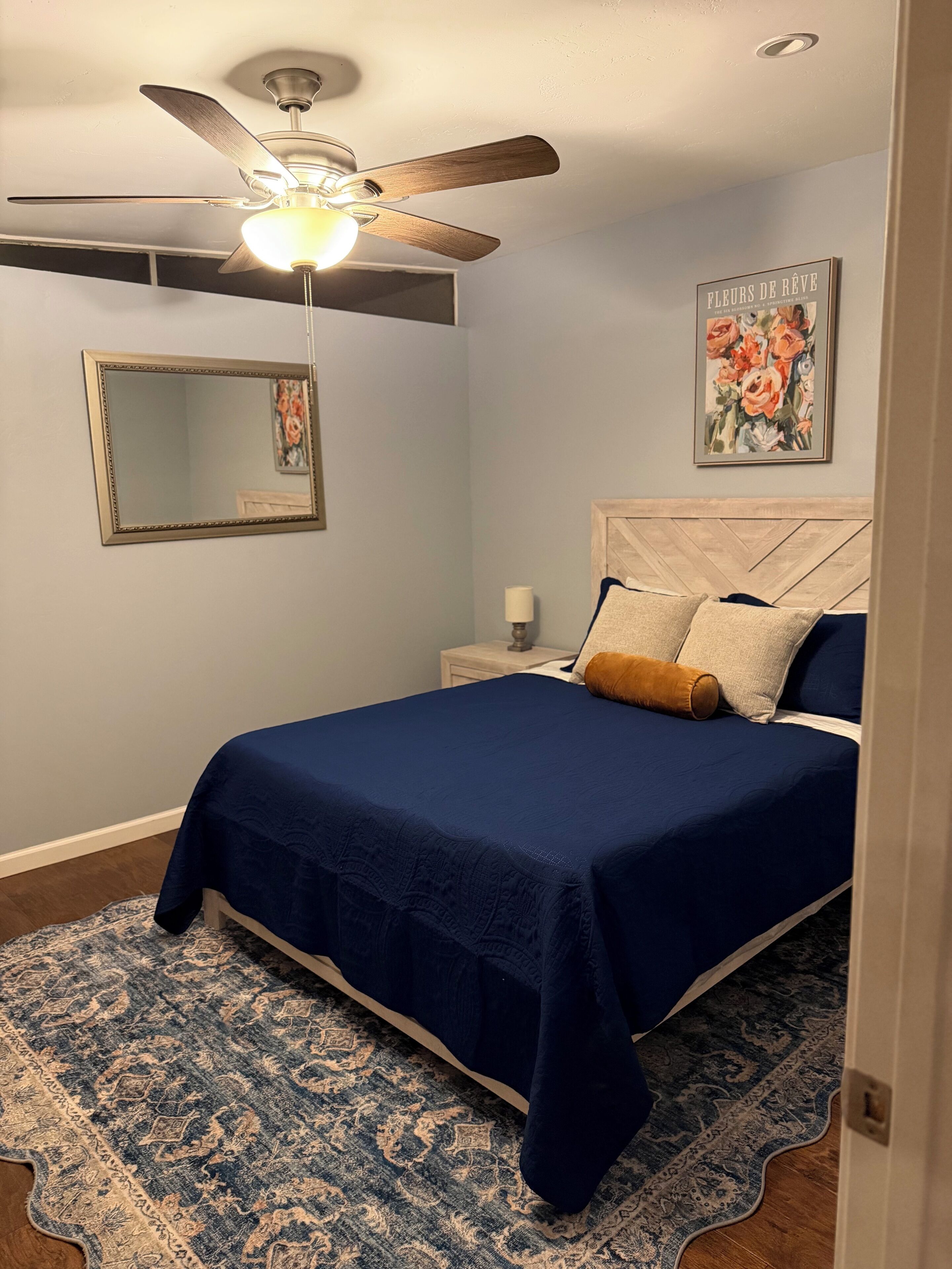 Quarto com cama, ventoinha no teto, mesa de cabeceira, espelho e um quadro na parede.