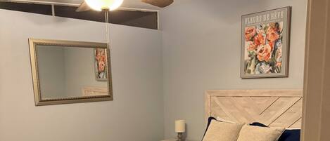 Quarto com cama, ventoinha no teto, mesa de cabeceira, espelho e um quadro na parede.