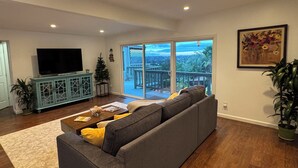 Ein Wohnzimmer mit einem großen grauen Sofa, einem Fernseher auf einer Kommode und einem Balkon mit Blick auf die Stadt.
