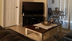 Ein Wohnzimmer mit Flachbildfernseher, einem dunkelbraunen Ledersofa, einem Couchtisch mit Untersetzer und einem Essbereich mit einer Glastür zum Balkon.