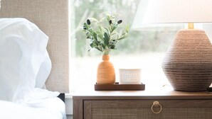 Un letto rifatto con lenzuola bianche, una lampada da comodino, un tavolino di legno con un vaso di foglie verdi e una candela.