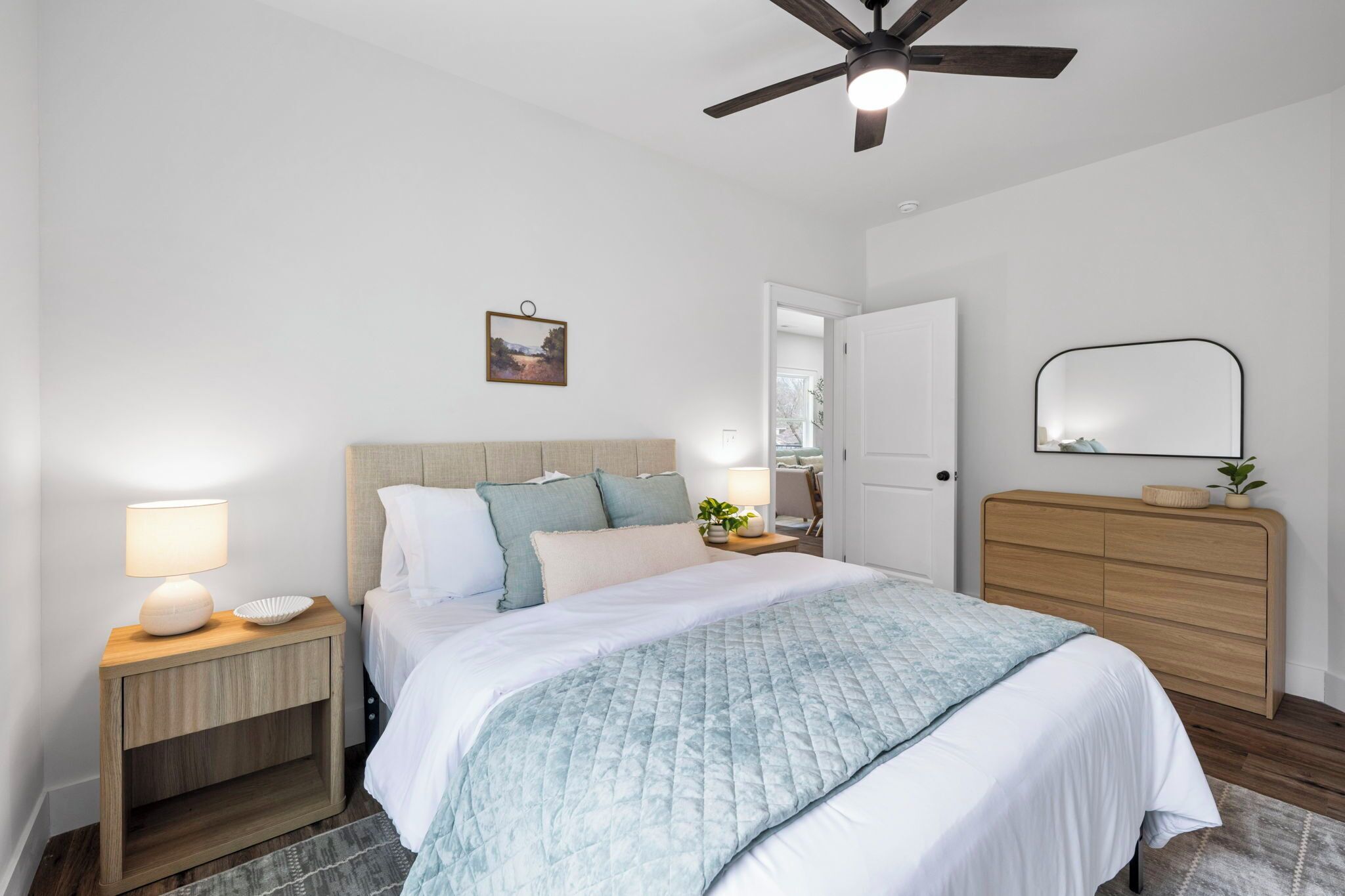 A bedroom with a bed, nightstands, a dresser, a ceiling fan, a door, and a wall-mounted mirror.