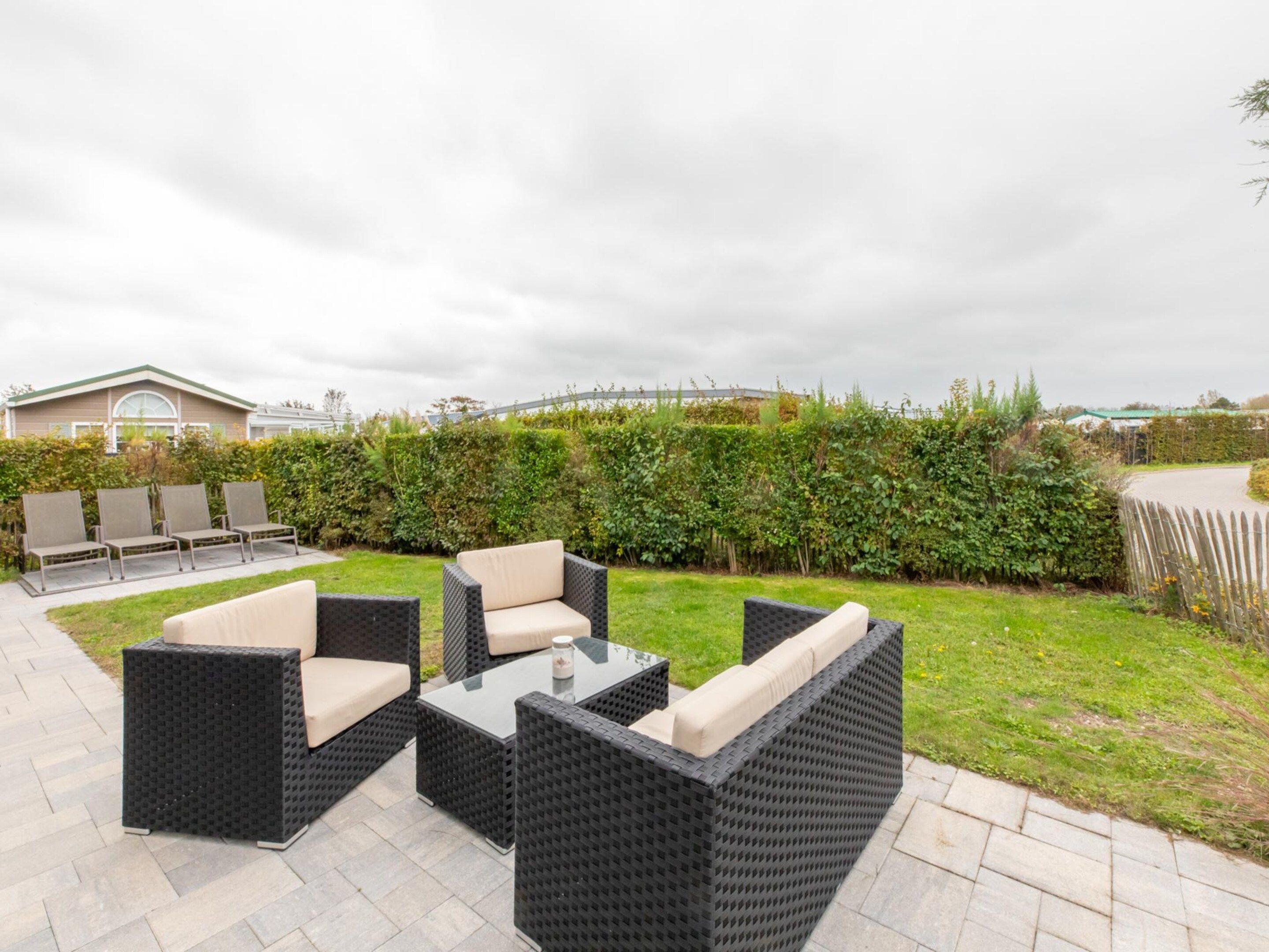 An outdoor seating area with wicker furniture and a table, surrounded by a hedge and a paved walkway.