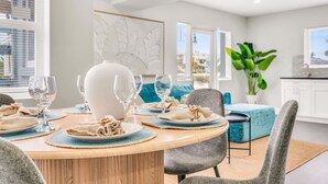 Ein Essbereich mit einem runden Tisch, gedeckt für vier Personen, umgeben von grauen Stühlen. Auf dem Tisch steht eine große weiße Vase, und der Raum hat ein modernes Design mit einem blauen Sofa und einer großen Zimmerpflanze im Hintergrund.