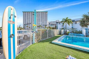 A pool area with a surfboard, a fenced deck, and a tall building in the background.