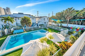 Une piscine entourée d’une clôture blanche, avec des palmiers et un bâtiment blanc en arrière-plan.