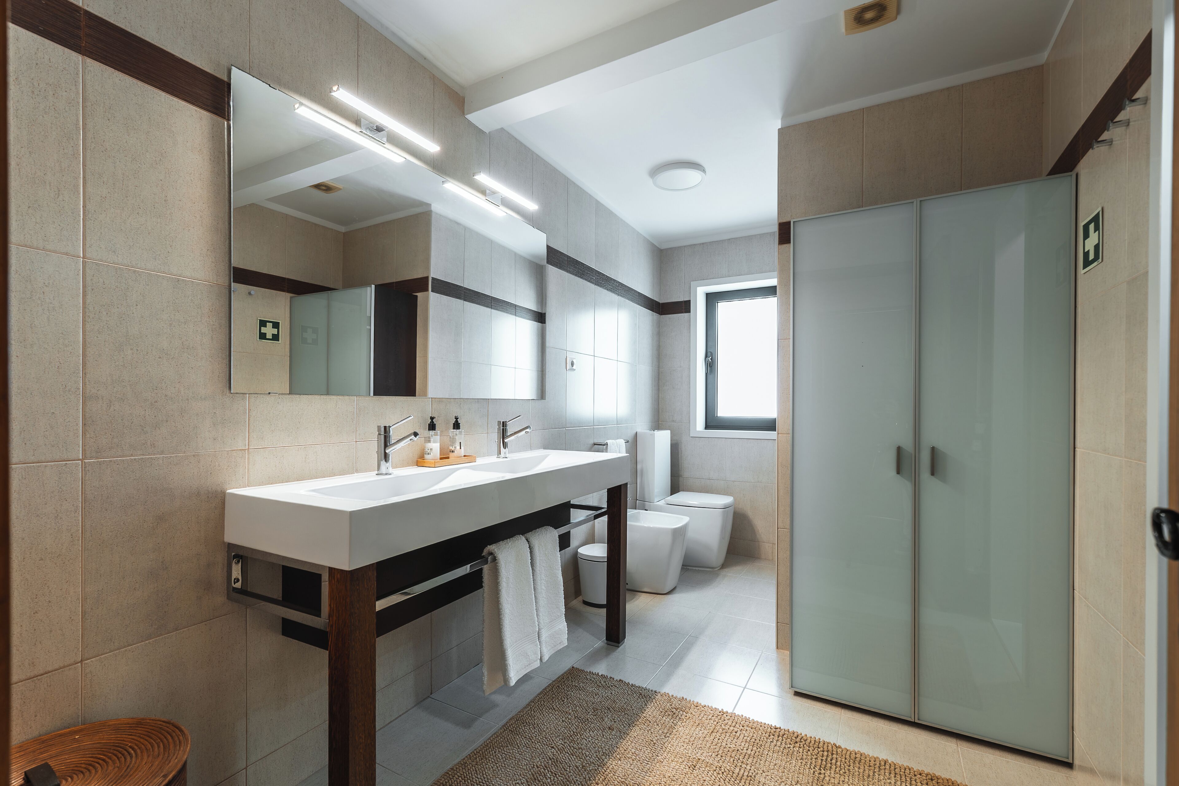 A modern bathroom with a large mirror, a white sink, a toilet, and a glass-enclosed wardrobe.