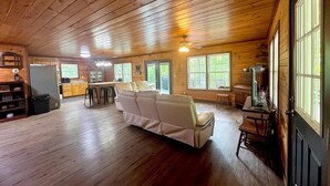 A spacious cabin with a large sofa, wooden floors, and a kitchen area. - Scenic 4‑BR sleeps 14 with Lake View, Decks, Private Dock, Kayaks, Canoe (LaFollette)