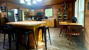 A wooden cabin kitchen with a central island, bar stools, a refrigerator, microwave, and a dining area with chairs. - Scenic 4‑BR sleeps 14 with Lake View, Decks, Private Dock, Kayaks, Canoe (LaFollette)