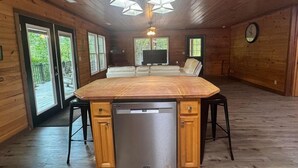 A wooden cabin interior with a central kitchen island, a dishwasher, and a seating area with a sofa and chairs. - Scenic 4‑BR sleeps 14 with Lake View, Decks, Private Dock, Kayaks, Canoe (LaFollette)
