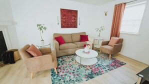 Een woonkamer met een beige bank, twee fauteuils, een salontafel en een kamerbreed tapijt. Twee potplanten, een rode decoratie aan de muur en een raam met gordijnen.