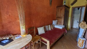 Ein rustikales Zimmer mit einem hölzernen Bett, einem kleinen Holztisch und einem Stuhl. Auf dem Tisch steht eine Bambuslampe und auf dem Boden ein Trommel.